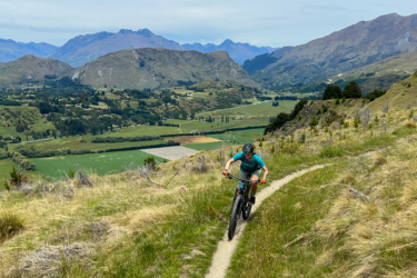 A person riding a mountain bike along a narrow trail on a hillside, with a scenic view of rolling hills, lush green fields, and distant mountains under a partly cloudy sky. Coronet Face Water Race mountain bike trail.
