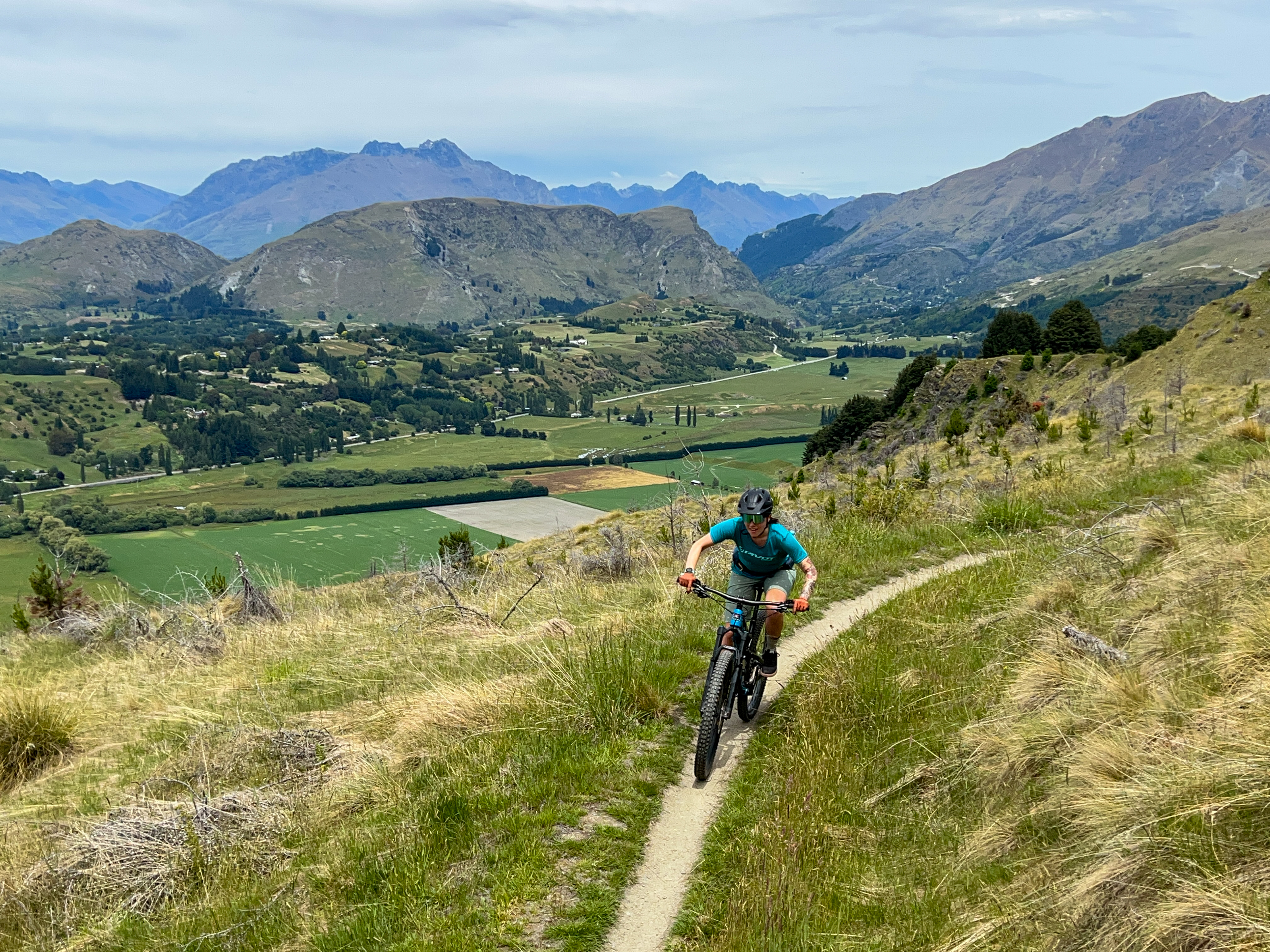 A person riding a mountain bike along a narrow trail on a hillside, with a scenic view of rolling hills, lush green fields, and distant mountains under a partly cloudy sky. Coronet Face Water Race mountain bike trail.
