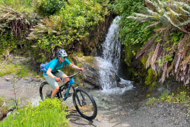 A person riding a mountain bike through a shallow stream in a lush forested area, with a small waterfall and ferns in the background. The cyclist is wearing a helmet and a turquoise shirt, navigating over rocky terrain. Coronet Face Water Race mountain bike trail.