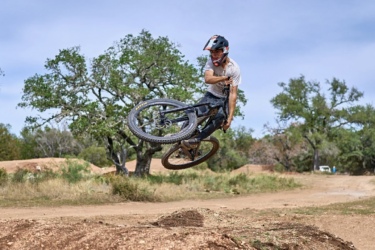 A mountain biker performing a trick in mid-air over a dirt jump, with a background of lush green trees and blue sky. The biker is wearing a helmet and casual clothing, showcasing dynamic movement and skill. Station Mountain Bike Park mountain bike trail.