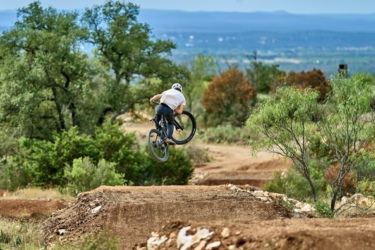 A mountain biker performing a jump over a dirt ramp in a natural setting, surrounded by trees and rolling hills in the background. Station Mountain Bike Park mountain bike trail.