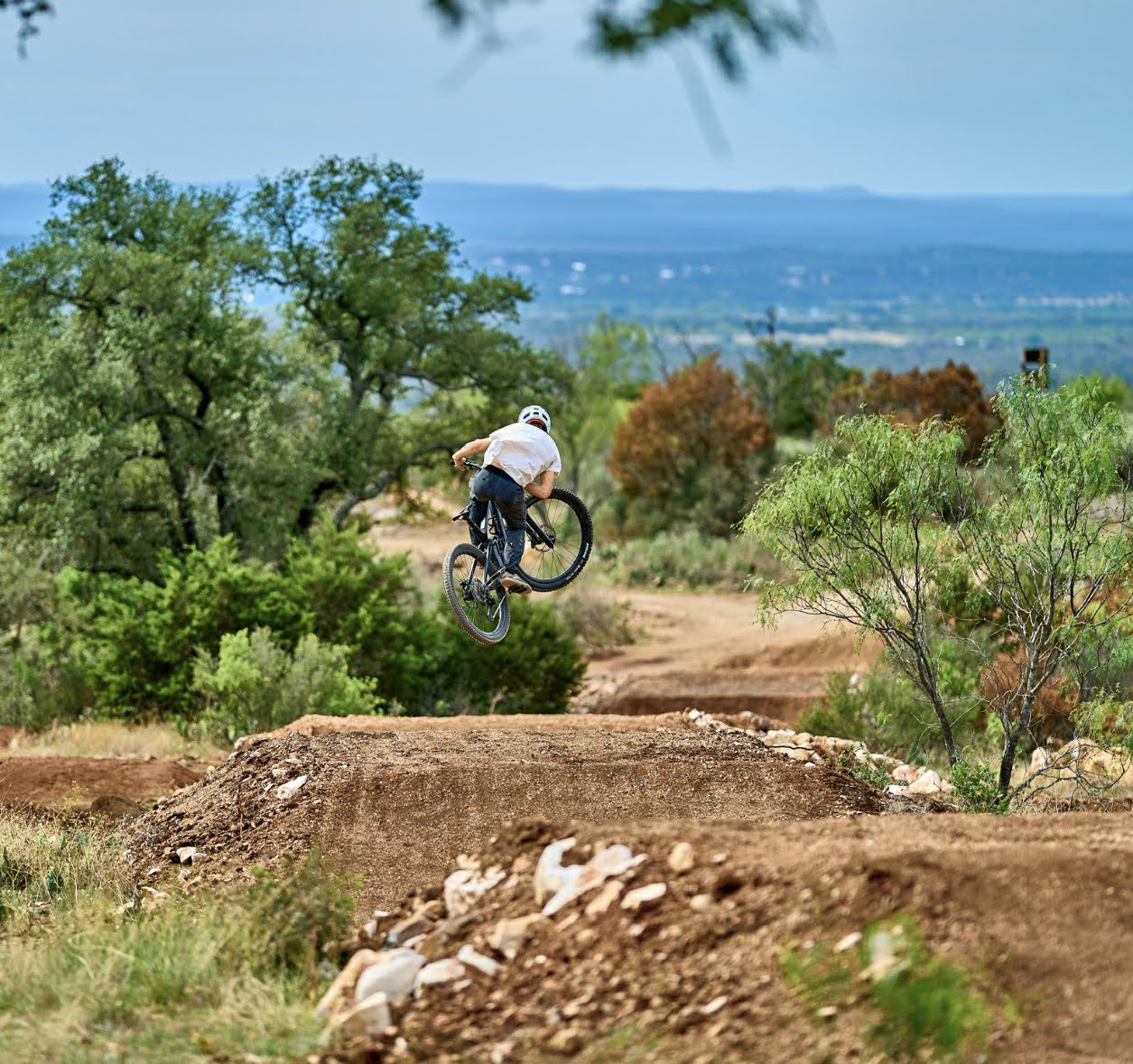 A mountain biker performing a jump over a dirt ramp in a natural setting, surrounded by trees and rolling hills in the background. Station Mountain Bike Park mountain bike trail.