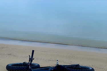 A fat-tire bicycle lying on its side on a sandy beach, with calm, turquoise waters and a cloudy sky in the background. The Beach mountain bike trail.