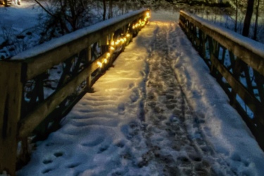 A snow-covered wooden bridge illuminated by warm lights, leading through a winter landscape at twilight. The footpath on the bridge shows signs of footsteps, and trees are silhouetted against a darkening sky. In the background, soft lights from nearby buildings hint at a peaceful winter scene. The Beach mountain bike trail.