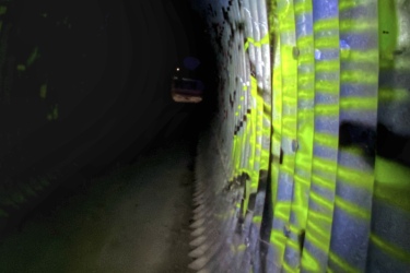 A dimly lit tunnel featuring curved metal walls with graffiti in shades of yellow and blue. The ground is visible, showing a concrete surface, and the tunnel extends into darkness in the distance. Light reflections create an interesting pattern along the walls. Rotary Memorial Trail mountain bike trail.
