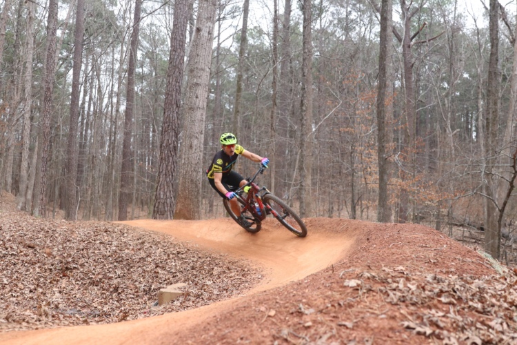 A mountain biker leans into a turn on a dirt trail surrounded by trees in a wooded area. The rider is wearing a bright yellow helmet and a black and yellow jersey, showcasing an energetic posture as they navigate the curve of the track. Fallen leaves cover the ground, adding to the natural setting.