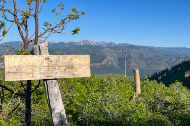 A wooden signpost reading "Hafen Creek" with an arrow pointing to the right, set against a backdrop of mountains and a clear blue sky. Green shrubs and trees are visible in the foreground, with a view of rugged mountain terrain in the distance. Haflin Creek mountain bike trail.