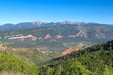 A panoramic view of a mountainous landscape featuring layered red rock formations, lush greenery, and a clear blue sky. The scene captures a mix of slopes and valleys, with distant snow-capped peaks appearing in the background. Haflin Creek mountain bike trail.