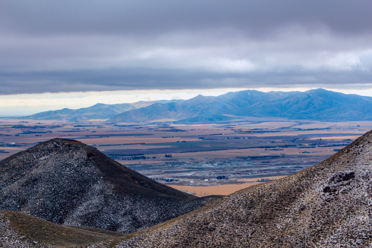 A panoramic view of rolling hills and mountains under a cloudy sky, with fields and agricultural land visible in the valley below. The foreground features rocky, snow-dusted hills, while the distant mountains are partially covered in a layer of snow.