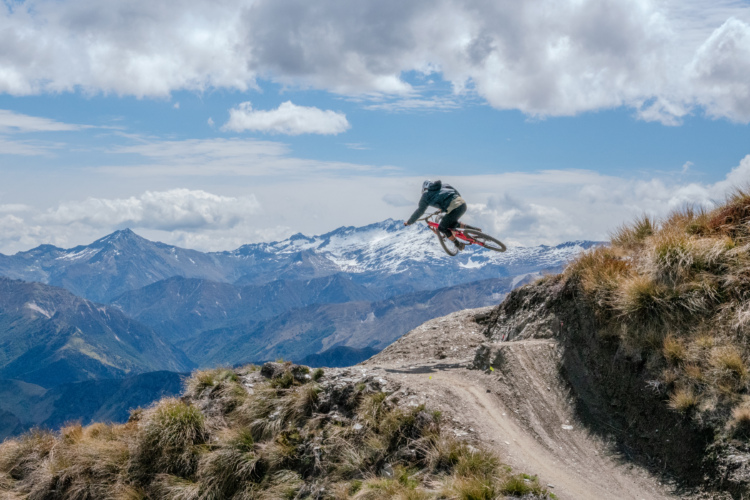 A mountain biker performing a jump on a dirt trail with a stunning backdrop of snow-capped mountains and a cloudy sky above.
