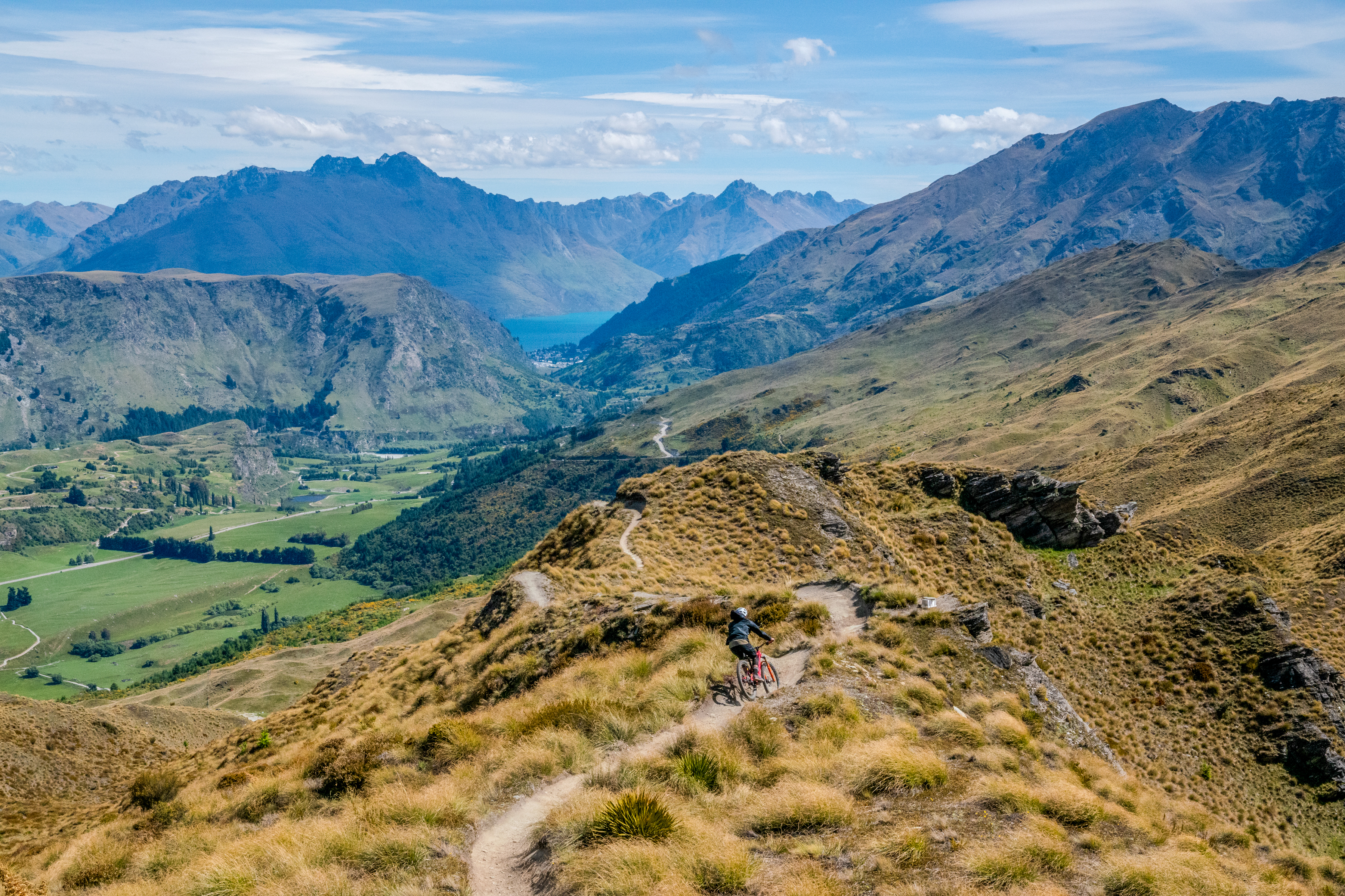 A mountain biker rides along a winding dirt trail on a grassy hillside, overlooking a lush valley and distant mountains under a clear blue sky. The scenery features rolling hills, patches of greenery, and a shimmering lake in the background. Rude Rock mountain bike trail.