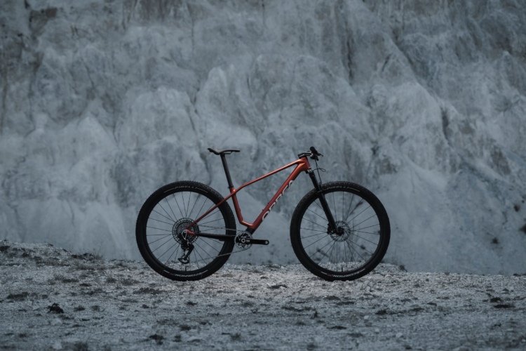 A modern mountain bike with a sleek orange frame stands on rocky terrain, set against a backdrop of textured, light-colored rock formations. The bike features thick tires and disc brakes, showcasing its off-road capabilities.