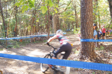 A mountain biker speeds around a dirt trail surrounded by trees, kicking up dust as they navigate the course. Blue safety tape is visible along the path, with spectators in the background watching the race. The scene captures the excitement of an outdoor cycling event in a forested area. The Ridgeland Trails mountain bike trail.