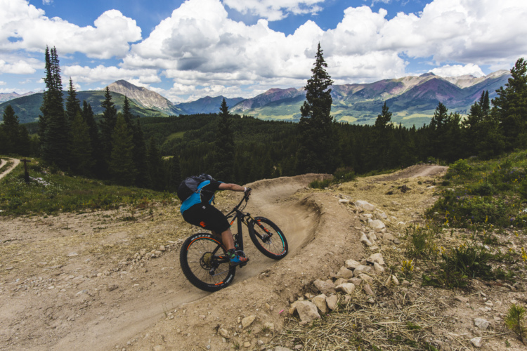 A mountain biker navigating a dirt trail with a curved turn, surrounded by lush green trees and expansive mountain views under a partly cloudy sky.