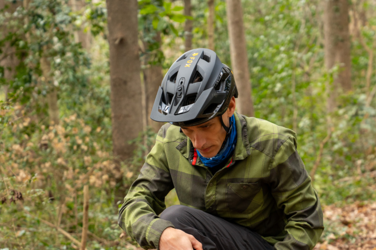 A person wearing a black cycling helmet and a green plaid shirt kneels on the ground in a forested area, surrounded by trees and greenery, with a focused expression as they engage with their surroundings.