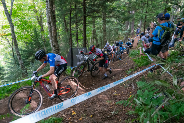 A group of mountain bikers navigating a steep, wooded trail during a race, with spectators cheering on the sidelines. The scene is set in a lush forest with greenery and a banner in the background indicating the location. Some riders are focused on their ascent while others are in different positions along the challenging course.