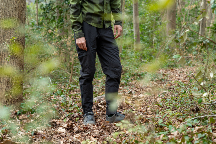 A close-up view of a person standing in a wooded area. The individual is wearing a green plaid shirt and Pearl Izumi Amfib Alpha insulated mountain bike pants. The ground is covered in fallen leaves, and greenery surrounds the scene, creating a natural, outdoor setting.