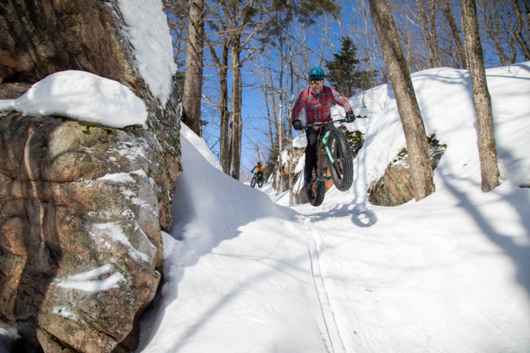 A fat bike rider performs a jump on a snowy trail, surrounded by trees and rocky terrain, under a clear blue sky. Another cyclist is visible in the background.
