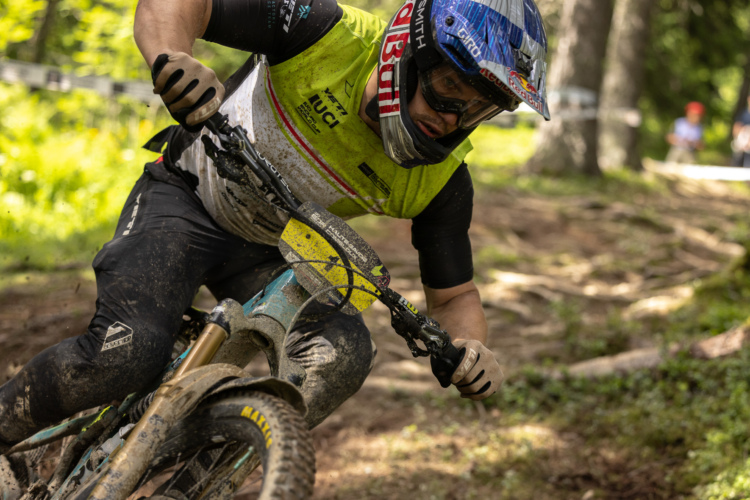 A mountain biker in a bright yellow and black jersey is leaning into a turn on a dirt trail, surrounded by trees. The cyclist is wearing protective gear, including a helmet and gloves, and their bike is partially obscured by mud, indicating an intense ride. Sunlight filters through the foliage, highlighting the action and the rugged terrain.