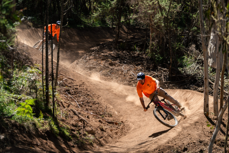Two mountain bikers in bright orange shirts navigate a dusty, winding dirt trail through a forested area. One rider leans into a turn while the other follows closely behind, both surrounded by tall trees and underbrush. Dust is kicked up behind them, capturing the dynamic energy of the activity.