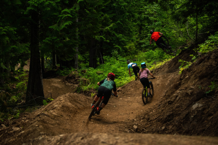 A group of young mountain bikers riding on a dirt trail in a lush green forest. One biker is airborne, performing a jump, while the others navigate the curves of the path. The scene captures the excitement and energy of outdoor biking activities.