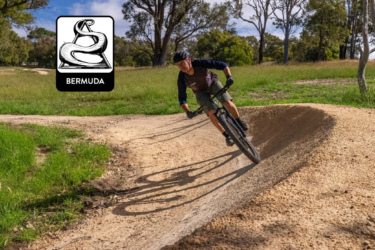 A mountain biker navigating a dirt track, leaning into a turn on a sunny day, with green grass and trees in the background. A logo featuring a stylized snake and the word "BERMUDA" is positioned in the corner of the image. Yalbunullup Mountain Bike Trails mountain bike trail.
