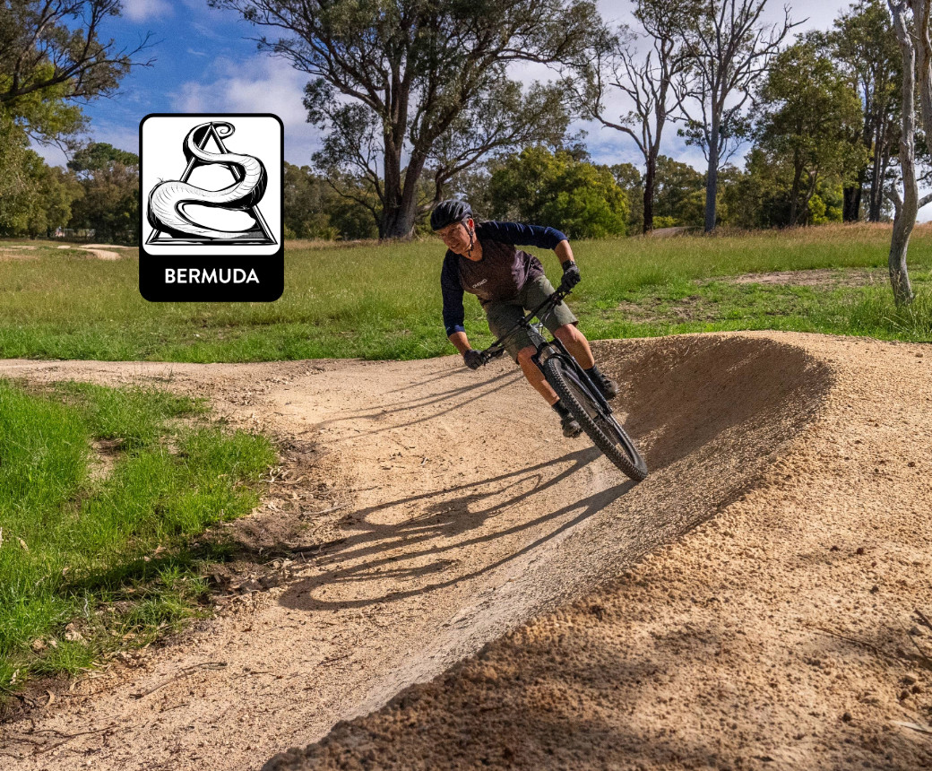 A mountain biker navigating a dirt track, leaning into a turn on a sunny day, with green grass and trees in the background. A logo featuring a stylized snake and the word "BERMUDA" is positioned in the corner of the image. Yalbunullup Mountain Bike Trails mountain bike trail.
