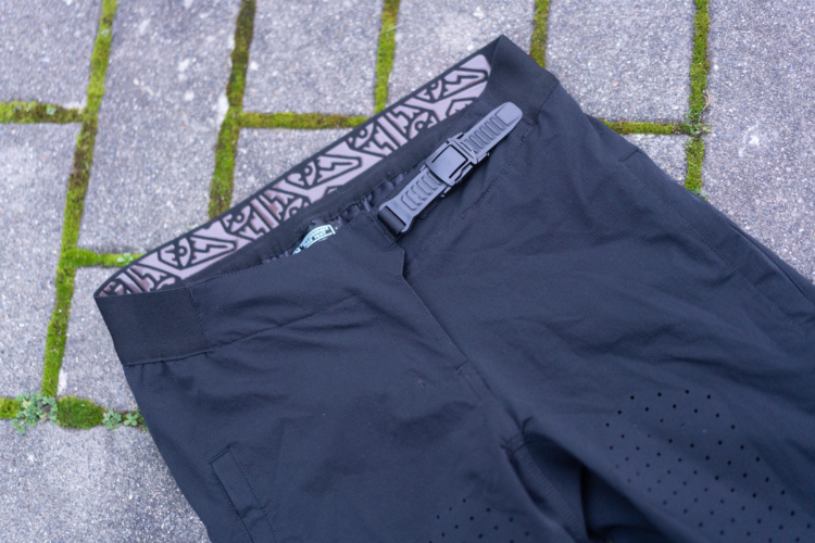 Black DFYRS Coffindodger pants laid flat on a textured stone surface, featuring a patterned waistband and an adjustable buckle. The pants have small perforations for ventilation along the sides.