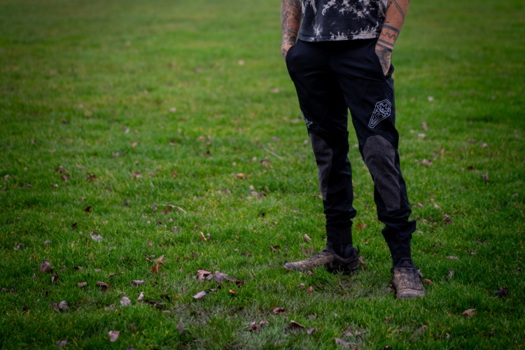 A person standing on a grassy field, wearing black DFYRS Coffindodger mountain bike pants with gray patches and a patterned black shirt. Their footwear is rugged and suitable for outdoor activity. The background features green grass with scattered leaves.