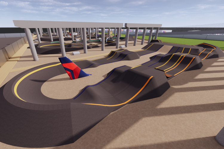 Aerial view of a modern skate park featuring a variety of ramps and obstacles. The surface is primarily dark with colorful lines marking the paths. The park is set under a structure supported by columns, with grassy areas visible in the background.