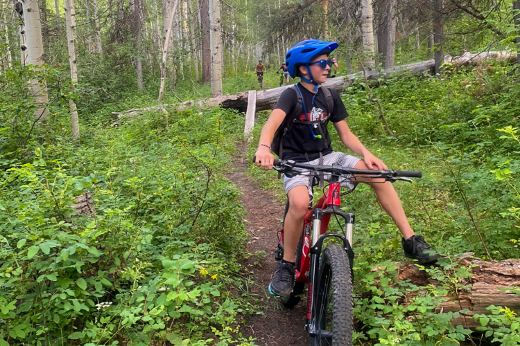 A young person wearing a blue helmet and sunglasses is sitting on a red mountain bike along a forested trail. Surrounded by lush greenery and tall trees, the rider appears to be enjoying a summer day outdoors, with a friend visible in the background.