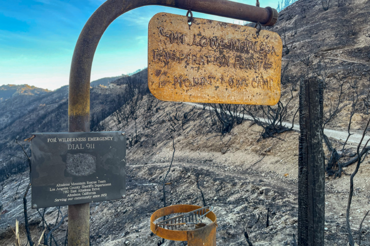 A weathered metal sign indicating "Lower Wilson Inspiration Point" hangs from a rusty pipe, alongside a plaque that instructs to dial 911 for wilderness emergencies. The background shows a landscape affected by wildfire, with charred earth and burnt vegetation. A winding path is visible in the distance, under a clear blue sky.
