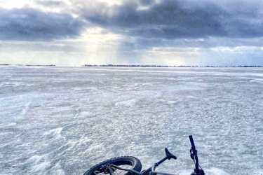 A fat bike lying on a frozen lake, with a cloudy sky and sunlight breaking through the clouds in the background. The icy surface reflects the overcast sky, creating a serene winter landscape. Rondeau Park mountain bike trail.