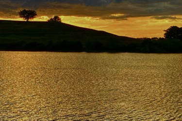A serene landscape featuring a calm lake reflecting a golden sunset. In the background, a gently sloping hill is silhouetted against the sky, with a few trees atop it. The sky is filled with dramatic clouds, merging shades of blue, orange, and gold, creating a peaceful and picturesque scene. Vista View Park mountain bike trail.