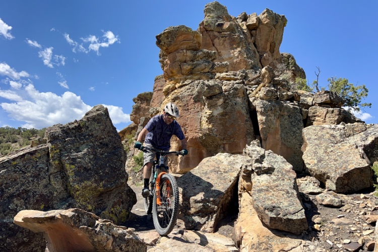 A mountain biker navigating a rocky trail surrounded by large boulders under a clear blue sky with scattered clouds.