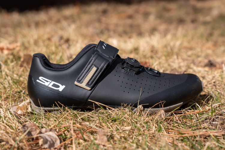 A close-up image of a sleek black cycling shoe, featuring a distinctive logo and a combination of straps and a dial for securing the fit. The shoe is positioned on grass with some fallen leaves, showcasing its smooth surface and perforated design for ventilation.