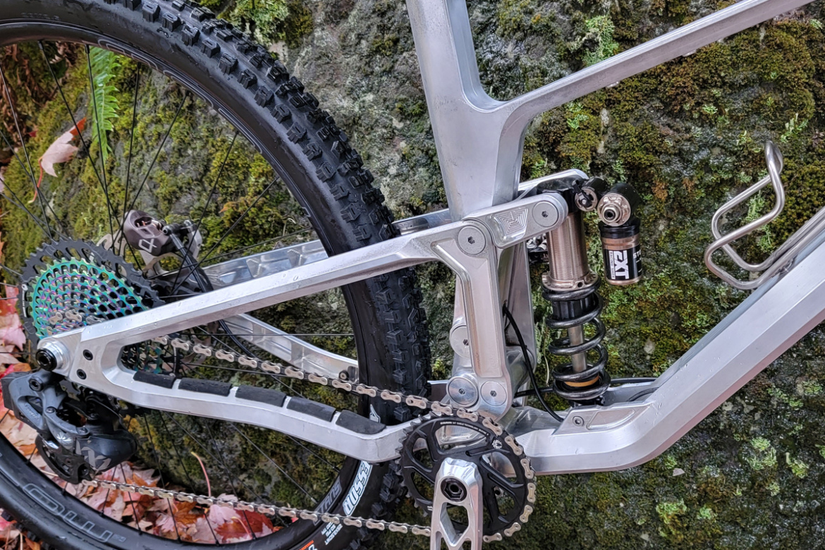 A close-up view of the rear suspension and drivetrain of a mountain bike, showcasing the intricate aluminum frame, chainring, and a colorful cassette. The background features moss-covered rock and fallen leaves, indicating an outdoor setting.