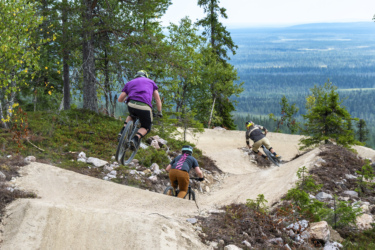 Three mountain bikers navigating a dirt trail in a forested area. The riders are shown from behind, with the first cyclist in a purple shirt airborne as they jump off a bump in the trail. The landscape features trees and a scenic view of rolling hills in the background. Yll&auml;s Bike Park mountain bike trail.