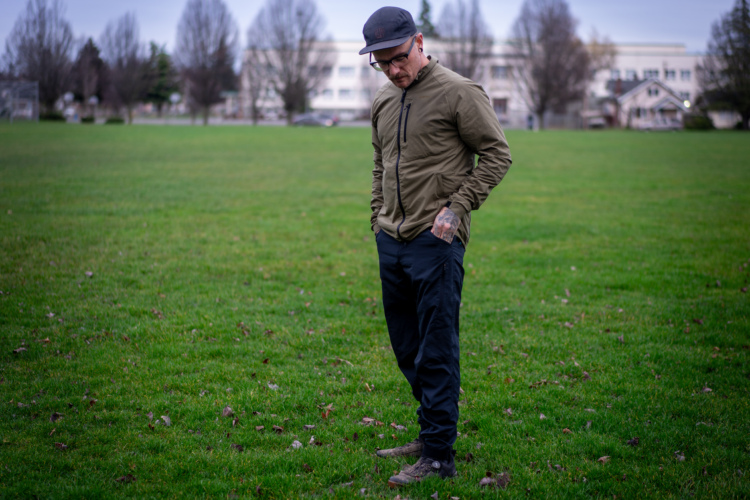 A person standing in a green park, looking down at the ground. They are wearing a Yeti Turq Edge jacket, black pants, and a cap. The background features bare trees and a building in the distance. The scene is set on a cloudy day.