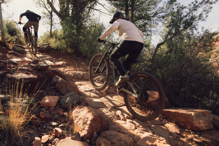 Two mountain bikers navigate a rocky trail surrounded by greenery. The first rider is ascending the path, showcasing a determined posture, while the second rider follows closely behind. The terrain is rugged with large stones and patches of dirt, under a clear sky.