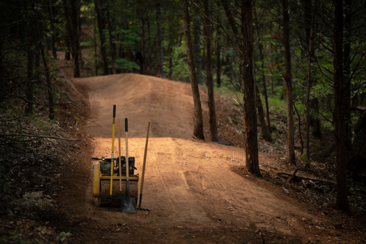 A collection of trail-building tools including a mini-tiller, a shovel, and a rake, resting on a dirt path in a wooded area. The path is gently sloped and surrounded by trees, with sunlight filtering through the foliage, illuminating the tools and the freshly prepared trail.