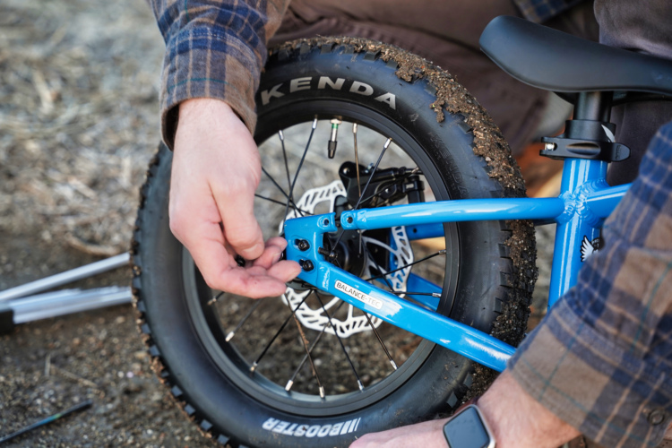 A person in a plaid shirt is adjusting the rear axle of a blue children's bicycle, focusing on a bolt near the large tire that has dirt on it. Tools are visible in the foreground on a gravel surface.