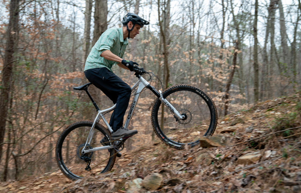 A cyclist wearing a helmet and gloves rides a mountain bike uphill on a rocky trail in a wooded area, with trees in the background and dry leaves scattered on the ground.