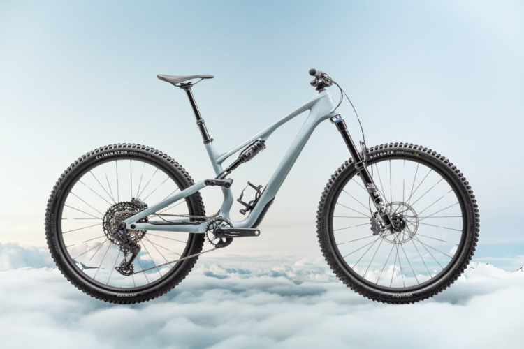 A sleek and modern mountain bike in a light grey color, positioned above a bed of white clouds against a soft blue sky. The bike features thick black tires and a detailed gear system, showcasing its design for off-road performance.
