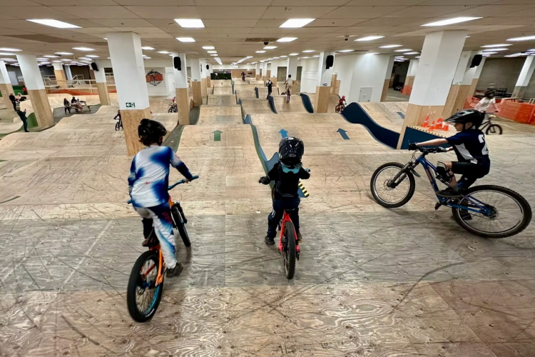 A group of children riding bicycles in an indoor bike park, featuring wooden ramps and obstacles. The scene shows four young riders in various riding positions, with two in the foreground wearing helmets. The park has a spacious layout with several jumps and turns, creating an active and energetic atmosphere.