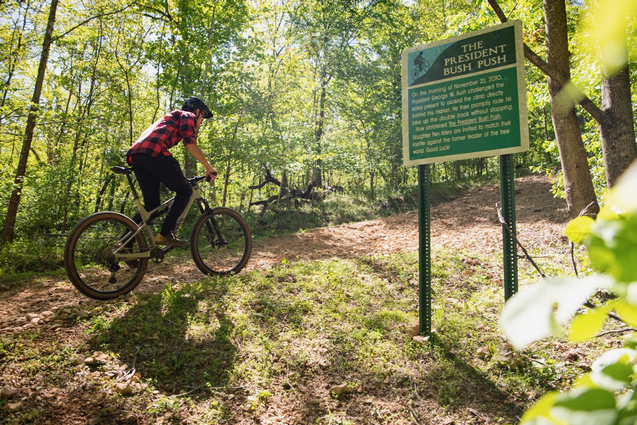A mountain biker wearing a black and red plaid shirt rides along a dirt trail in a wooded area, alongside a sign titled "The President Bush Push," which describes a challenge related to biking. Sunlight filters through the trees, casting dappled shadows on the trail. President Bush Push mountain bike trail.