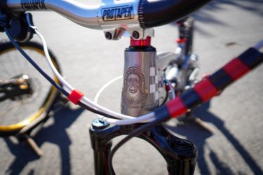 Close-up view of a bicycle's handlebars and stem, featuring a silver headset with a decorative logo, colorful cables, and a black fork. The background shows a blurred view of the bicycle wheels on a paved surface.