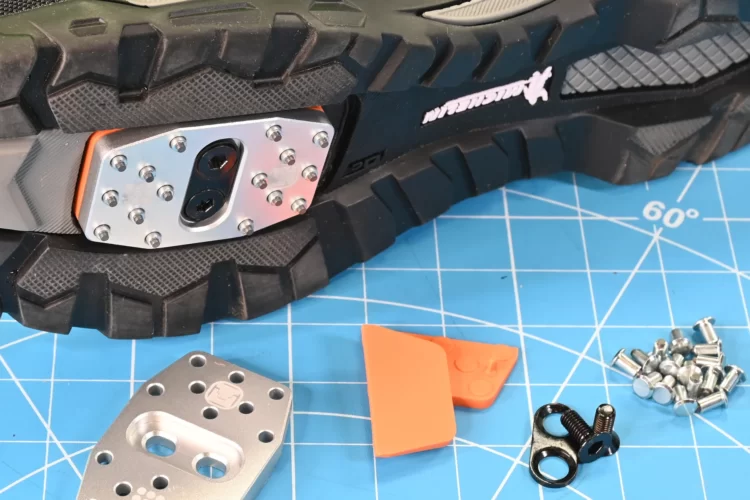Close-up of a hiking shoe's outsole featuring a detachable metal plate with holes for attaching equipment, an orange fitting tool, and loose hardware components, set against a blue background with a grid pattern.