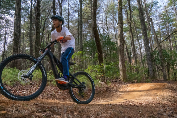 A young rider in a helmet balances on a mountain bike while navigating a dirt trail in a wooded area, surrounded by tall trees and greenery.