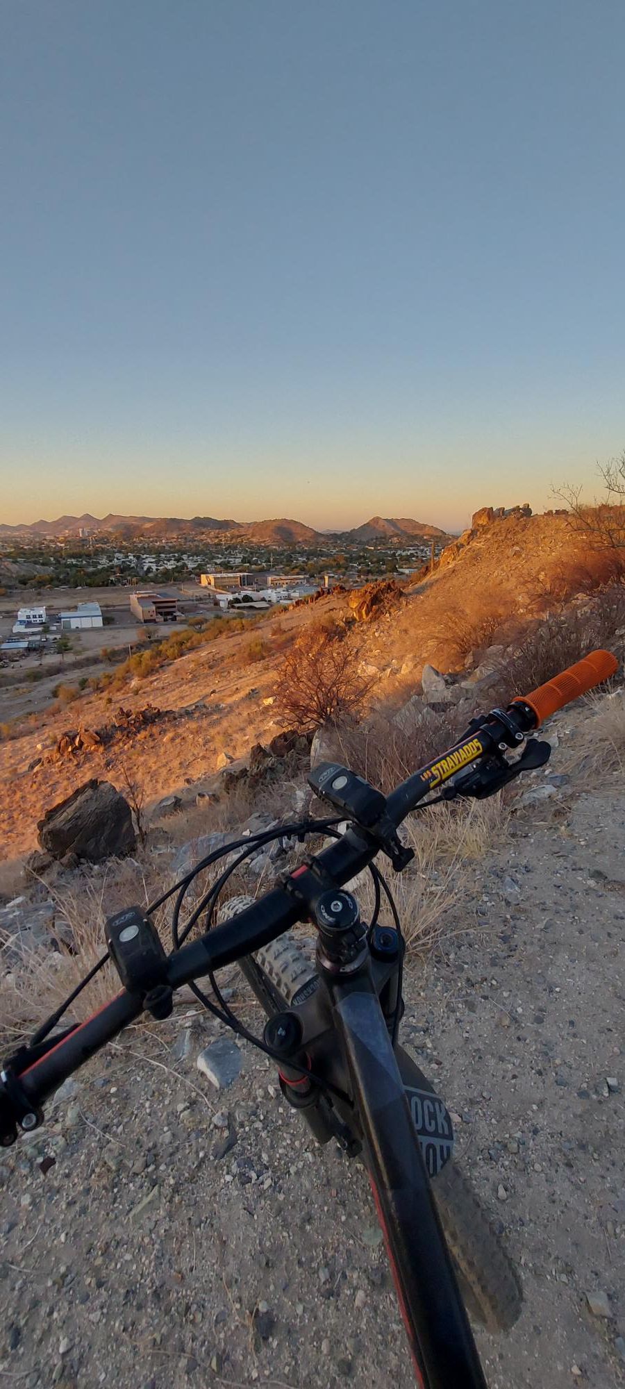 Alt text: A mountain bike rests on a rocky trail with a scenic view of a valley and mountains in the background during sunset. The bike